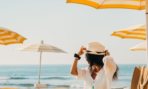 Persona in spiaggia con cappello bianco, ombrelloni sullo sfondo.