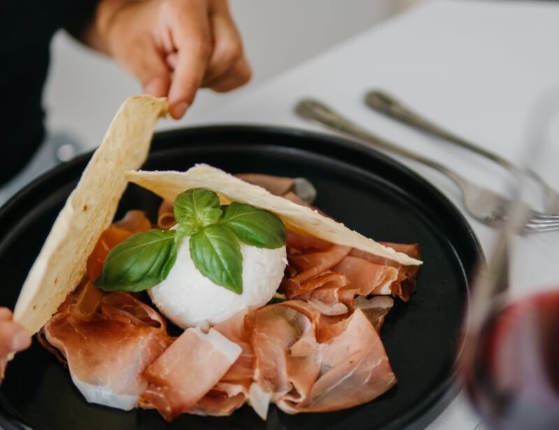 Piatto nero con salumi, formaggio, basilico, e pane. Vino rosso sfocato.
