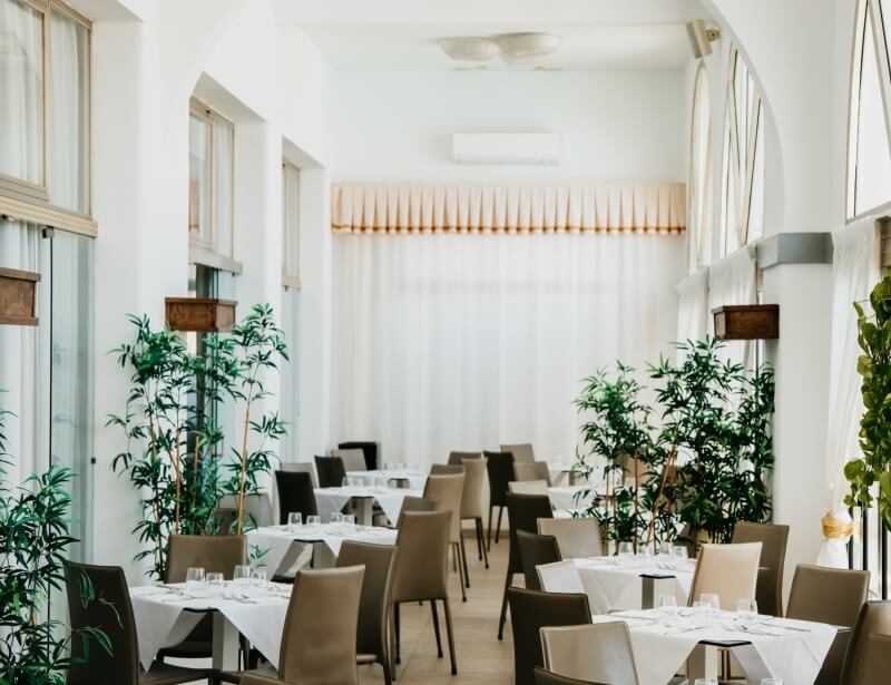 Area ristorante con tavoli bianchi, piante e tende bianche.