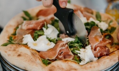 Pizza with prosciutto, cheese, and greens being cut with a cutter.
