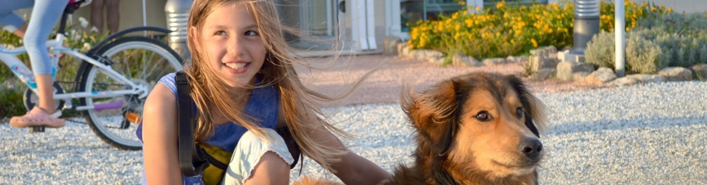 Ein Mädchen mit blonden Haaren kniet auf Kies neben einem Hund, während ein Fahrrad und ein Garten im Hintergrund sichtbar sind.