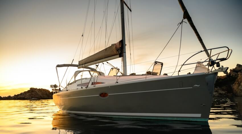 Vela su acqua calma al tramonto, barca grigia con vela avvolta, riflessi del cielo giallo sull'acqua.