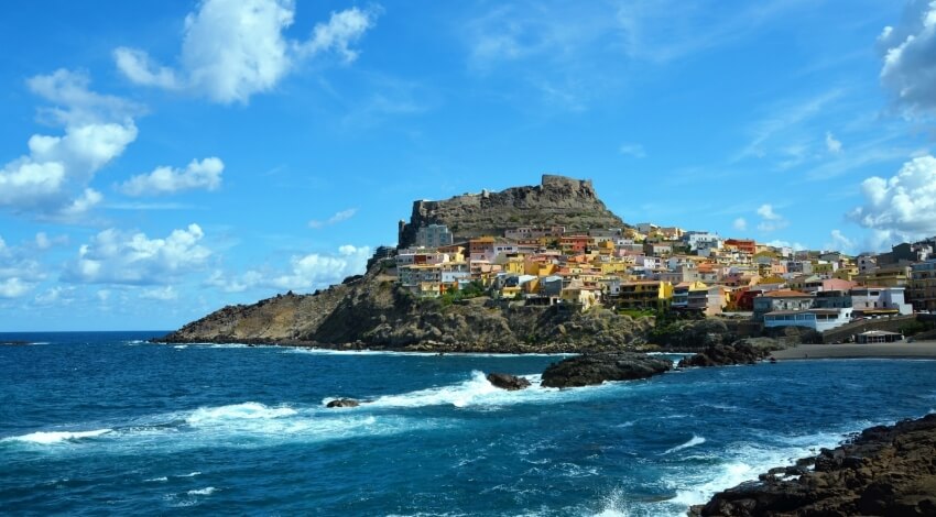 Una pittoresca cittadina costiera con edifici bianchi e gialli, un colle roccioso e un mare blu che increspano le onde.