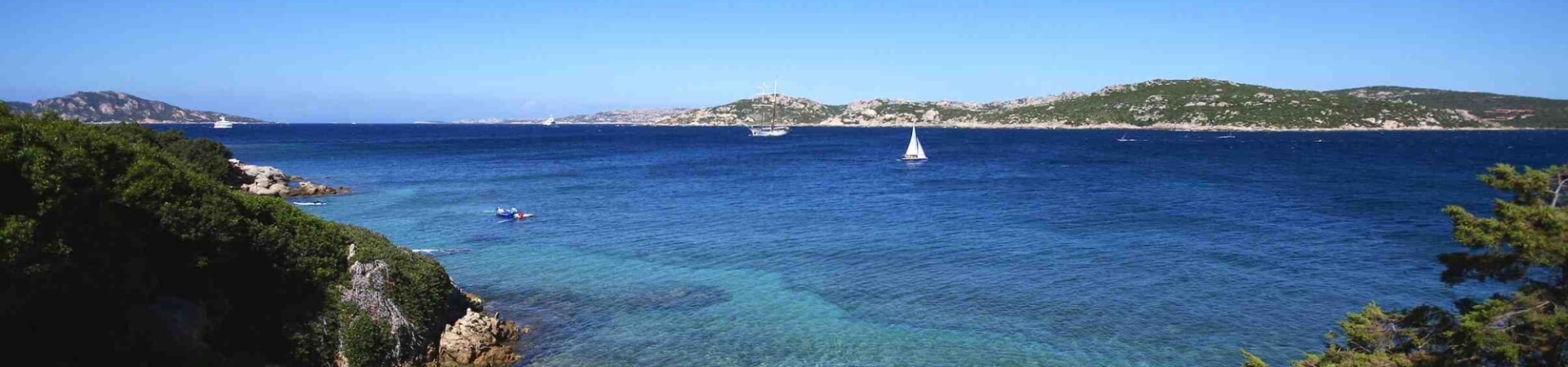 Una scena costiera serena con acqua calma, barche e kayak, circondata da vegetazione e una costa rocciosa all'orizzonte.