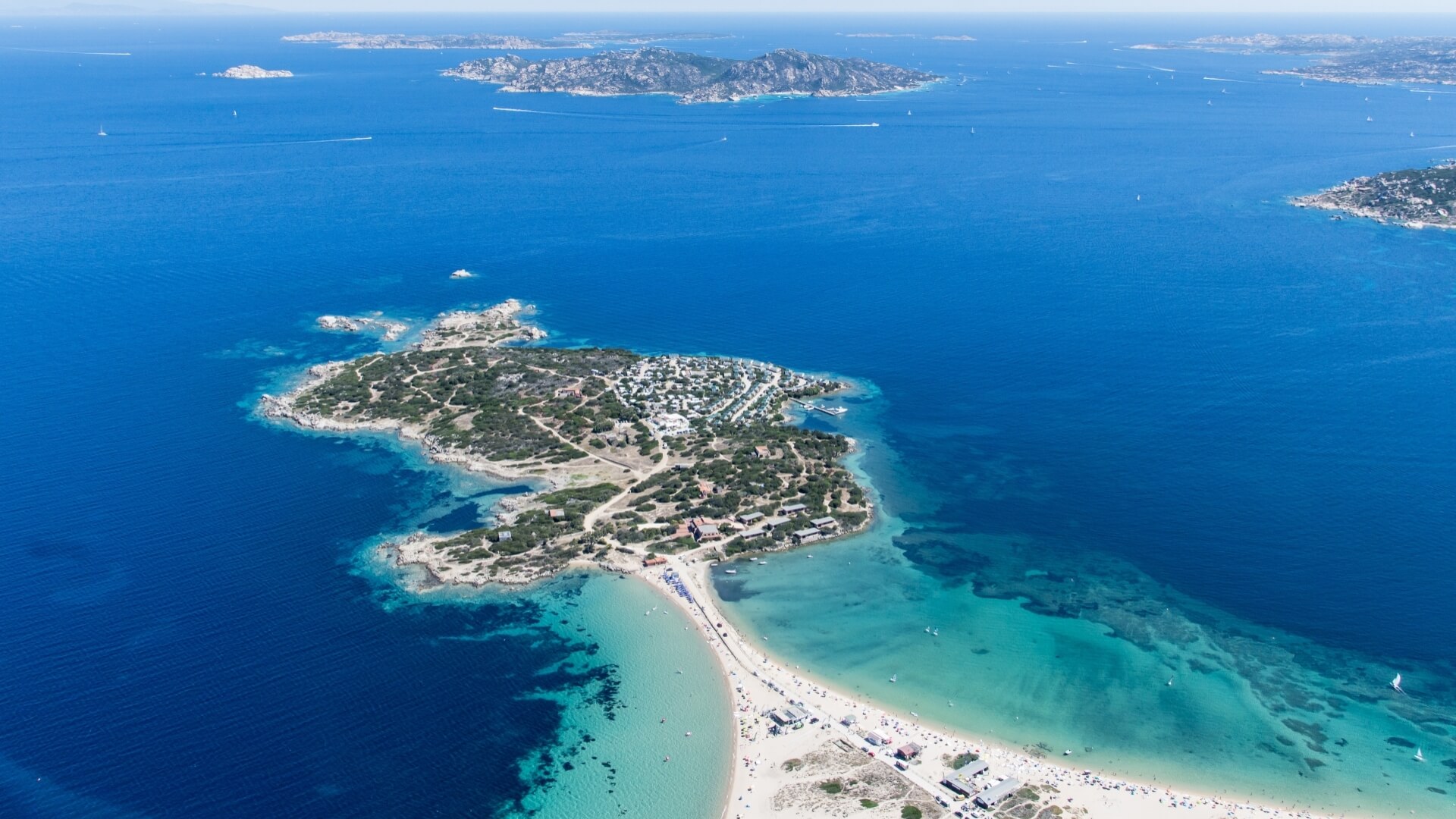 Vista aerea di un'isola serena circondata da acqua blu, con spiaggia, persone e isole più piccole sullo sfondo.