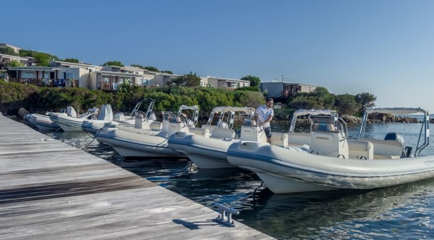Marina serena con un molo in legno e cinque barche bianche, un uomo in camicia bianca è sulla seconda barca da destra.