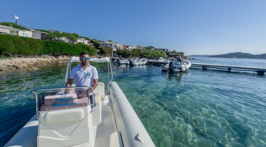 Una barca bianca naviga in acque turchesi, con un uomo al timone, verso una costa pittoresca.