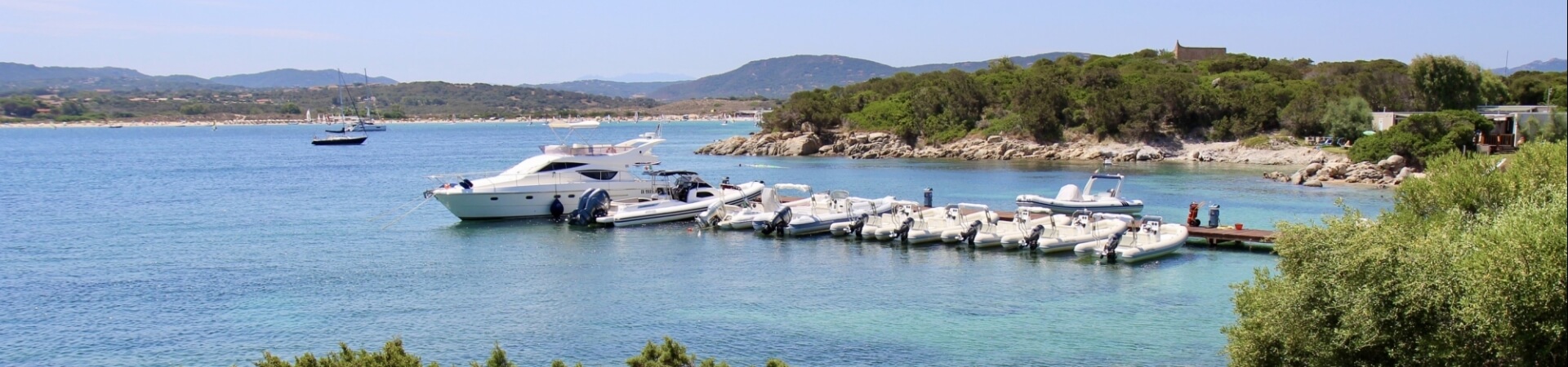 Serene coastal scene with calm water, moored boats, lush greenery, and rolling hills under a clear blue sky.