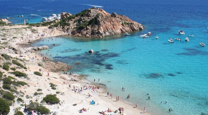 Ein ruhiger Strand mit klarem Wasser, Felsen im Hintergrund, Menschen am Strand und Boote im Wasser.