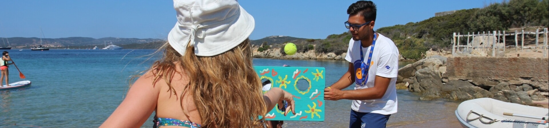 A woman in a bikini plays a game with a man on the beach, while another woman paddleboards in the background.