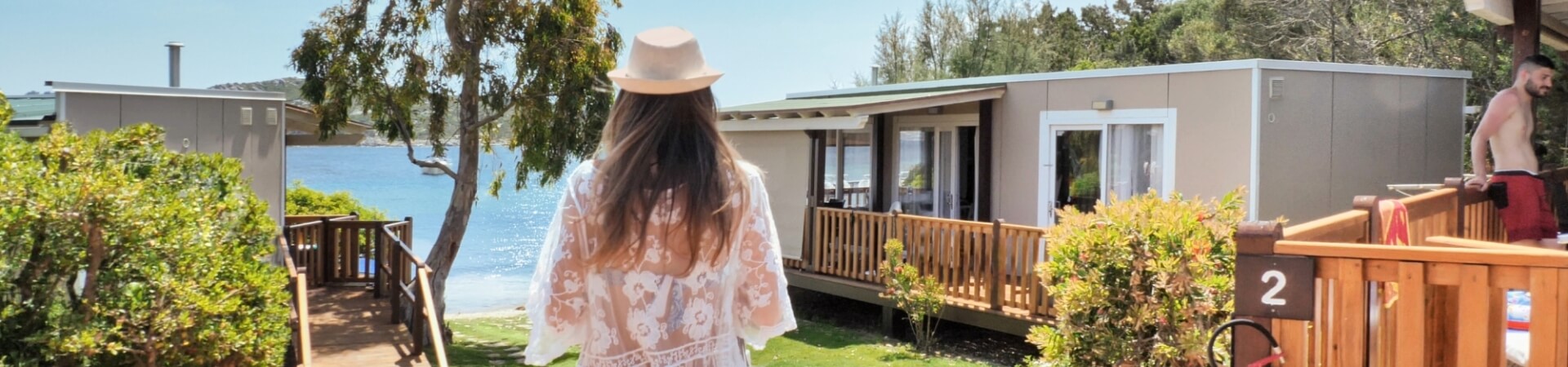 Eine Frau in einem weißen Kleid und Hut steht auf der Veranda eines Wohnmobils, während ein Mann in roten Badehosen auf einem Deck lehnt.