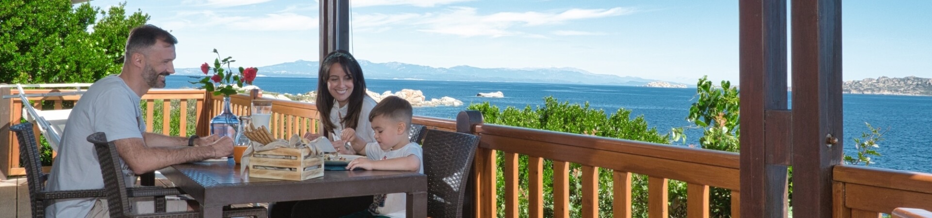 Una famiglia di tre persone cena su un deck con vista sull'oceano, circondata da alberi e un'atmosfera serena.