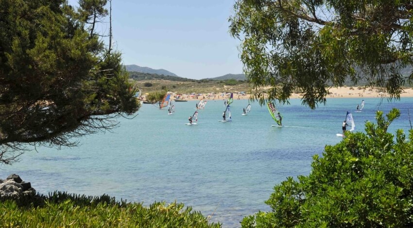 Persone windsurfing in una baia con spiaggia e colline verdi.