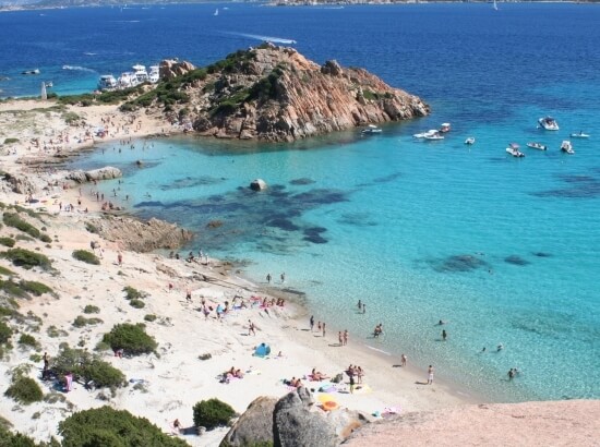 Serene beach scene with rocky hill, turquoise waters, sandy shores, sunbathers, boats, and distant buildings under blue skies.