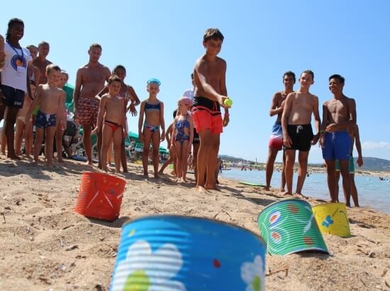 A group of children plays on the beach with cans, while a boy prepares to throw a green ball and a lifeguard watches.