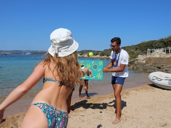 A man and woman play a beach game while others prepare for activities, under clear skies and calm waters.