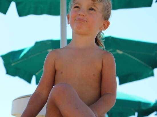 A young child sits cross-legged in the sun, gazing upward, with three green umbrellas providing shade behind them.