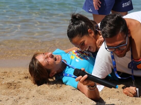 A woman lies on the beach with a microphone, assisted by a man; a staff member stands in the water behind them.