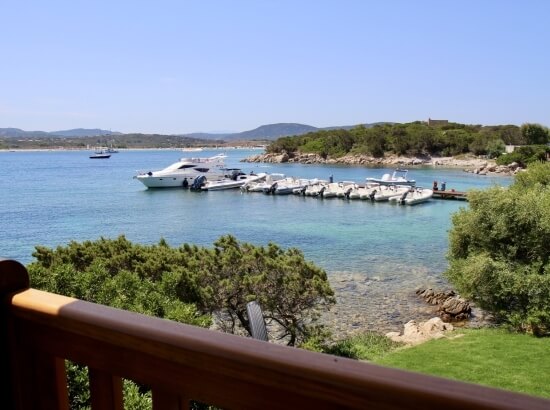 Serene waterfront scene with a wooden railing, calm water, boats, a distant dock, rocks, and lush trees on the shore.