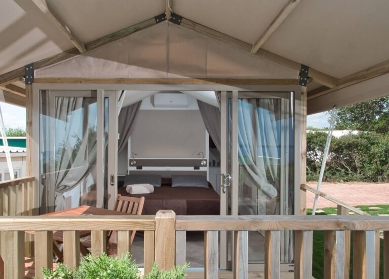 Tenda glamping moderna con interni accoglienti, patio esterno, piante verdi e vista su alberi e cielo blu.