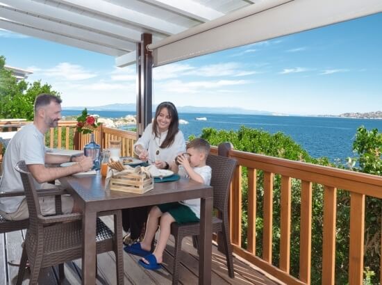 Una famiglia si gode un pasto su un deck, con vista su un corpo d'acqua sereno, sotto una struttura di ombra.