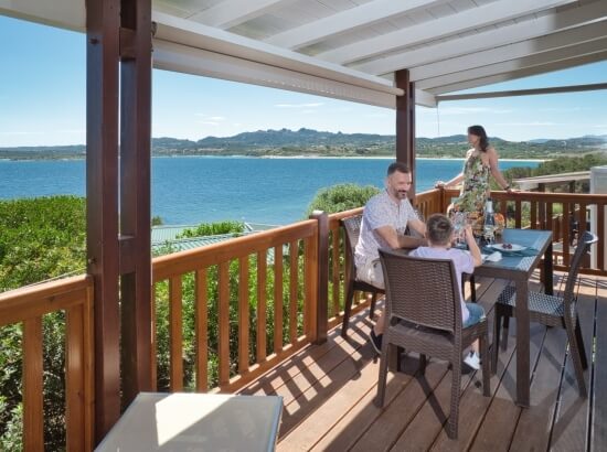 Eine Familie genießt eine Mahlzeit auf einer Holzterrasse mit Blick auf einen ruhigen See und eine bergige Landschaft.