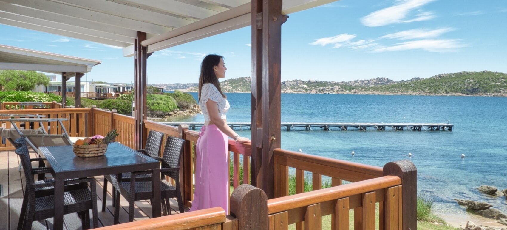 Una donna con capelli scuri in piedi su un balcone, guarda un corpo d'acqua sereno circondato da vegetazione.