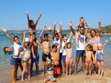 Gruppo di 18 persone in spiaggia, mani alzate, con acqua chiara a sinistra e alberi a destra, cielo blu sopra.