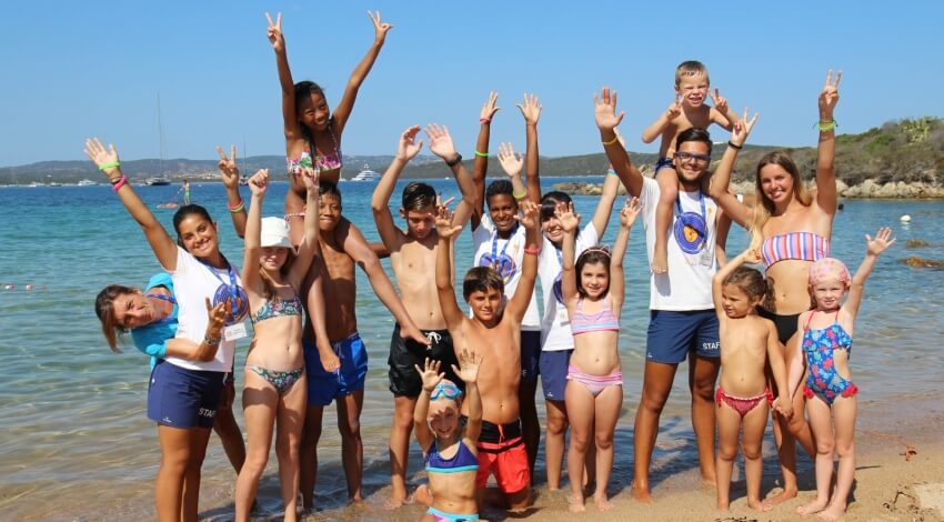 Un gruppo di 17 persone, tra cui bambini e adulti, posa sulla spiaggia con le mani alzate e un cielo blu sullo sfondo.