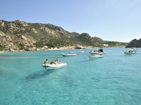 Boats in light turquoise water near a rocky coastline under a cloudy sky, evoking serenity and tranquility.