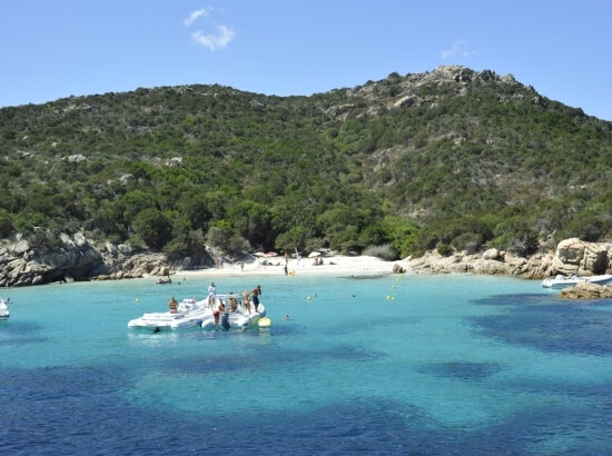 Serene coastal scene with a turquoise bay, forested hill, and people near the water's edge with a paddle boat.