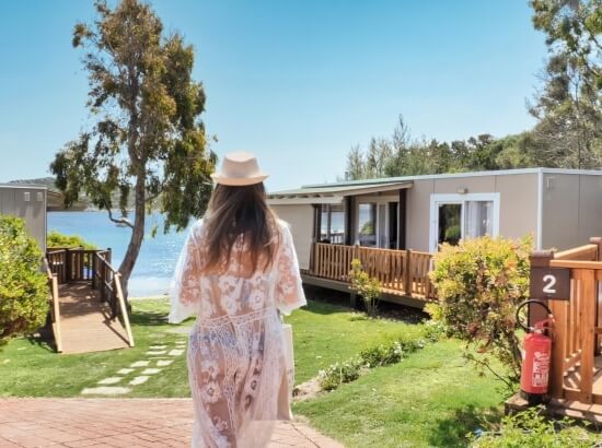 Eine Frau mit einem weißen Hut und Bademantel geht zu einem Haus am Meer unter einem klaren blauen Himmel.