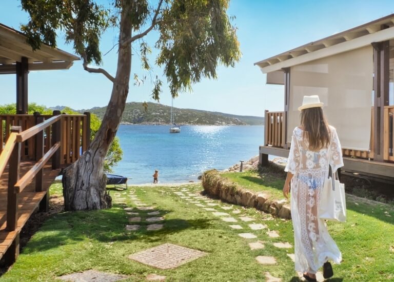 A woman in a white dress walks towards the water, with buildings, a child on the beach, and hills under a blue sky.
