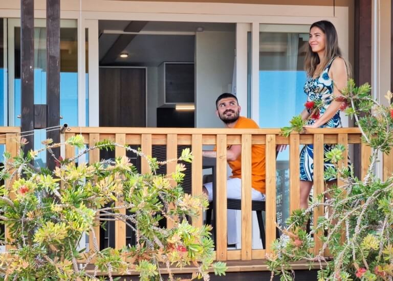 Ein Paar steht auf einem Balkon mit Holzgeländer, umgeben von üppigem Grün und roten Blumen, mit einer Glasschiebetür im Hintergrund.