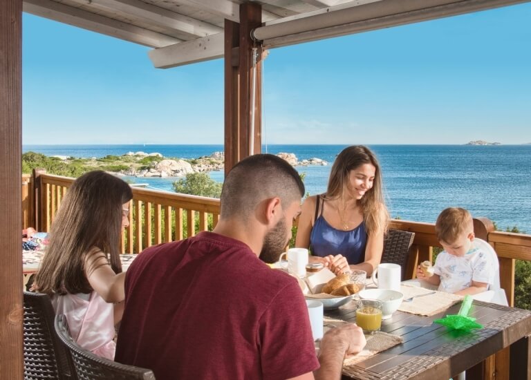 A family of four enjoys breakfast on a balcony with an ocean view, seated at a wooden table with food and drinks.