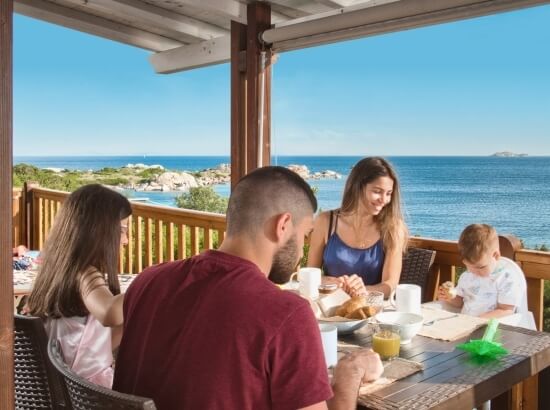 Una famiglia di cinque persone fa colazione su un patio con vista sull'oceano, circondato da un'atmosfera serena.