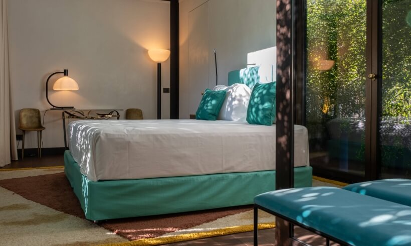 Modern bedroom with a bed, teal accents, nightstand, chair, and natural light from a window.