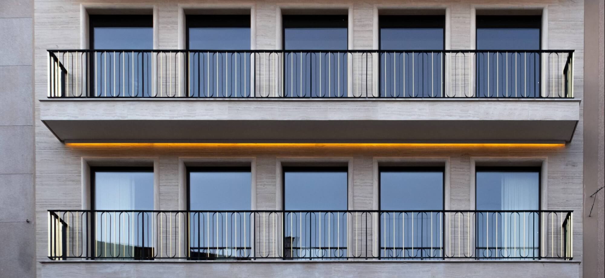 Modern building with two levels of balconies, black railings, light-colored stone exterior, and yellow light beneath the top balcony.