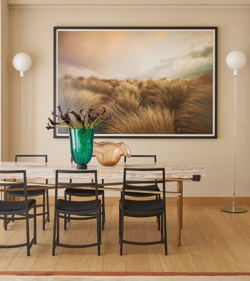 A serene dining room with a light wood table, black chairs, purple flowers, and artwork of golden grass on beige walls.
