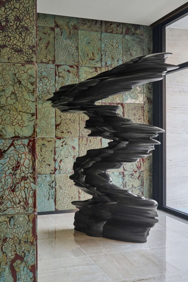 Abstract dark gray sculpture resembling waves, surrounded by green and blue tiled walls and a light beige tiled floor.