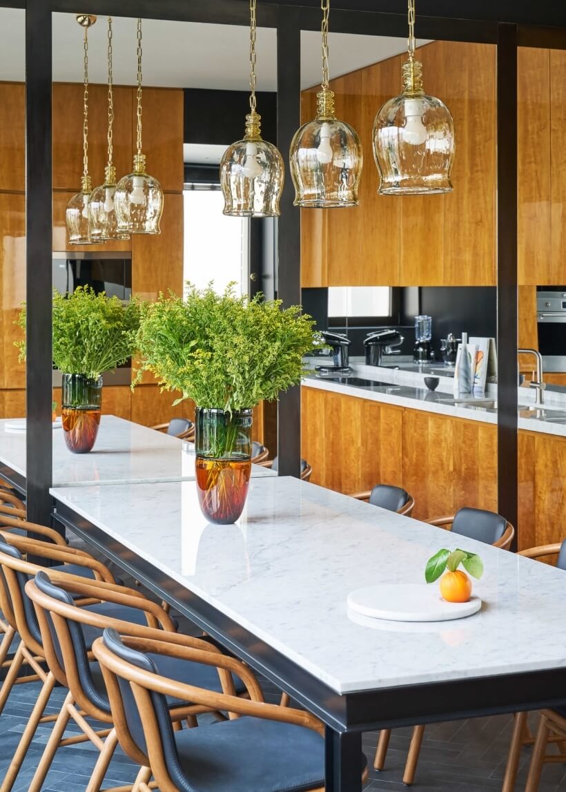 Modern kitchen with a marble dining table, greenery vases, pendant lights, and sleek wooden cabinets in an elegant setting.