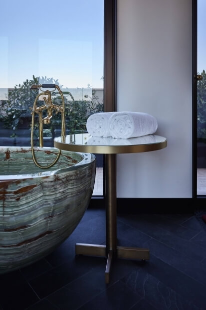 Luxurious bathroom with a freestanding marble tub, gold faucet, a round table with towels, and large windows showcasing a serene view.