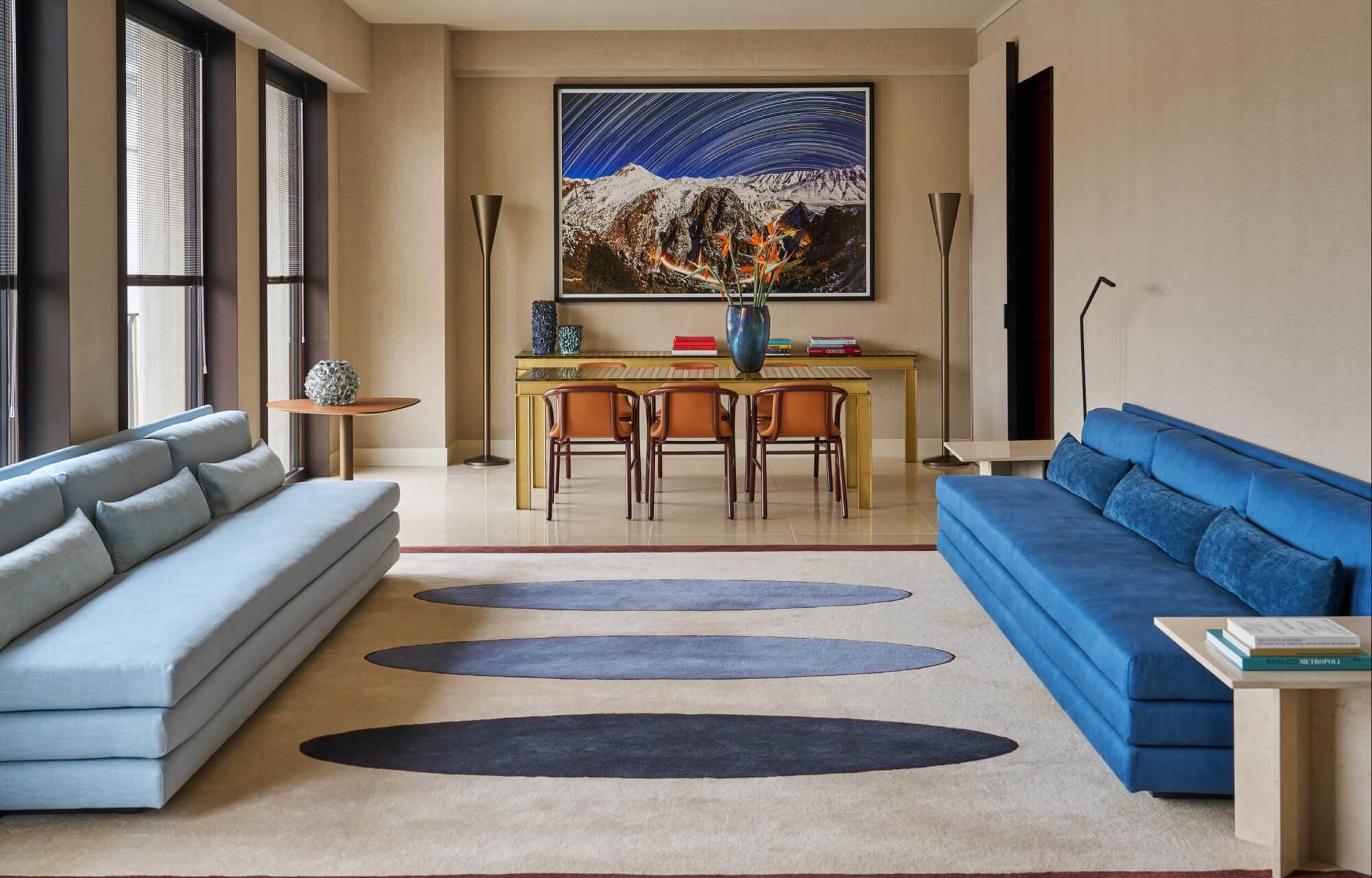 Modern living room with two blue sofas, wooden dining table, large landscape painting, and natural light from windows.