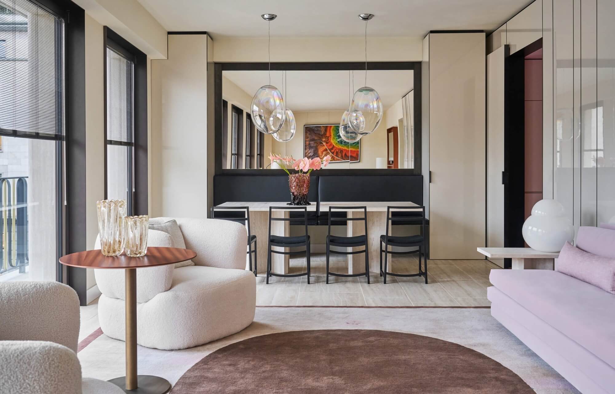 Modern living room with dining area, large windows, pink couch, and elegant decor, featuring a mirror and pendant lights.