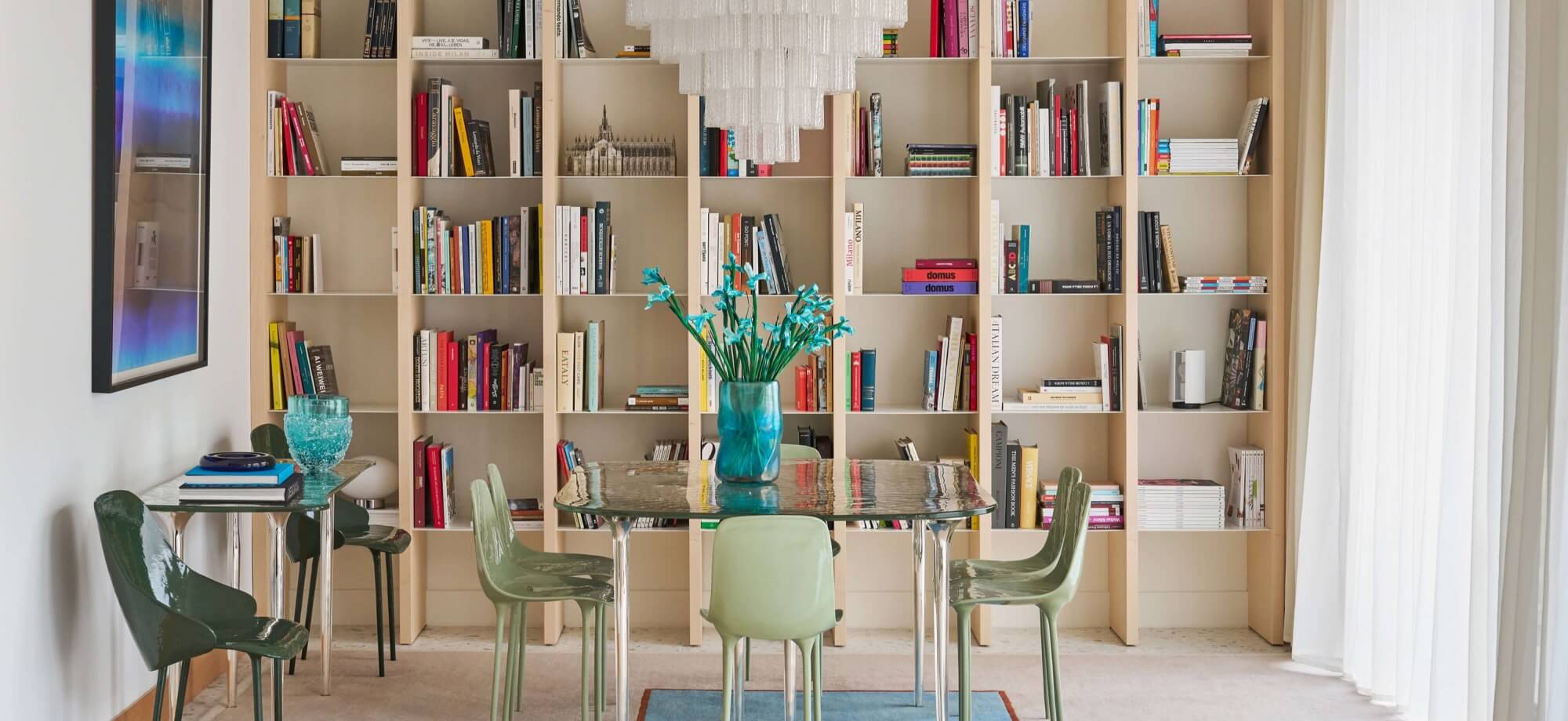 A mid-century modern dining room with a glass table, green chairs, a large bookshelf, and a small table with blue flowers.