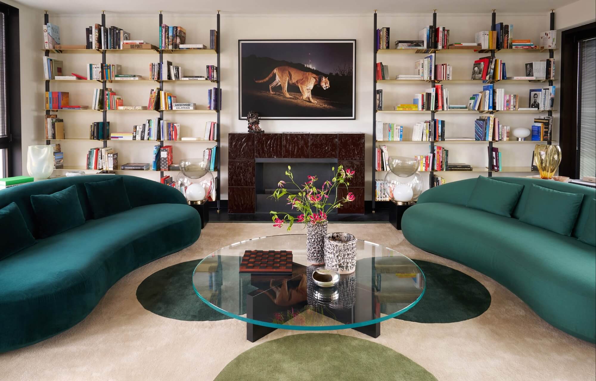 Modern living room with teal couches, a fireplace, bookshelves, and a glass coffee table adorned with flowers and decor.