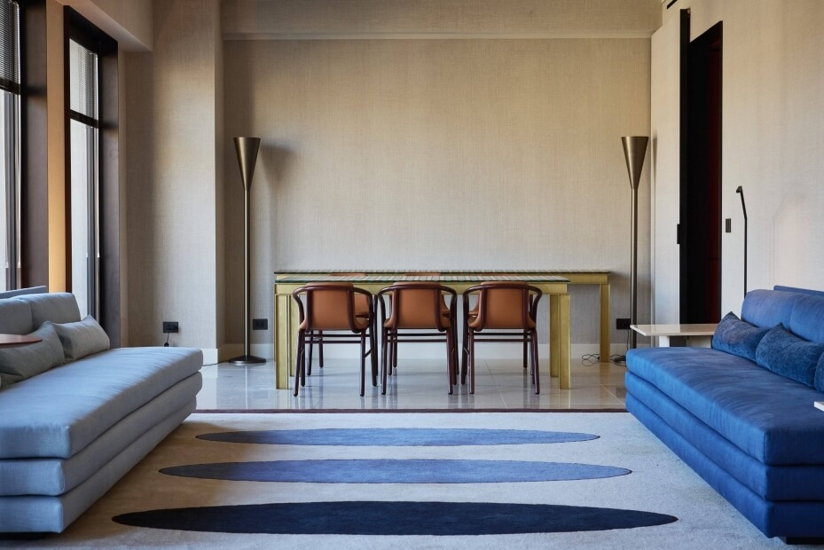Modern living room with large windows, light gray couch, blue couch, wooden table, and uniquely designed rug.