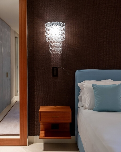 A dark-brown wall with a white light fixture, open doorway to a light gray room, and a bed with blue and white bedding.