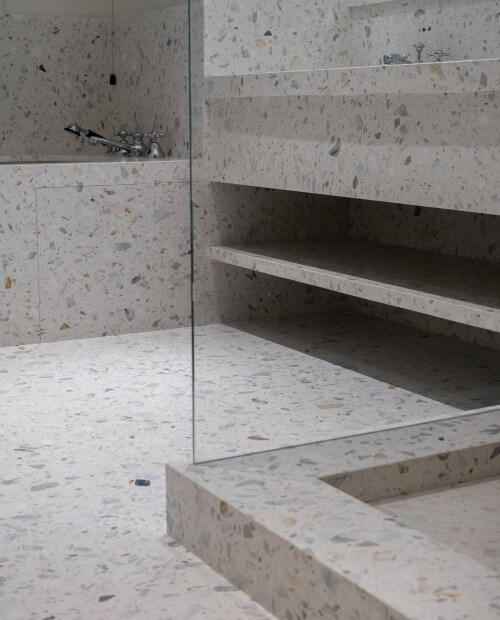 Modern bathroom with terrazzo floor and walls, featuring a shower with a silver faucet and a storage shelf.