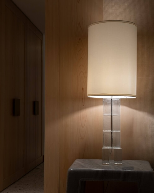 A lit lamp with a white shade and clear glass base on a nightstand, casting a warm glow against a wood-paneled wall.